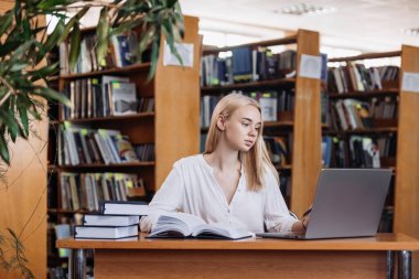 Ciddi derecede sıkı çalışan sarışın kız öğrenci webinar izliyor, çevrimiçi öğrenim kurslarını dinliyor, dizüstü bilgisayara bakıyor, üniversite kütüphanesindeki masada notlar yazıyor.