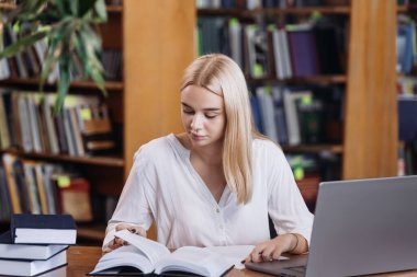Ciddi derecede sıkı çalışan sarışın kız öğrenci webinar izliyor, çevrimiçi öğrenim kurslarını dinliyor, dizüstü bilgisayara bakıyor, üniversite kütüphanesindeki masada notlar yazıyor.