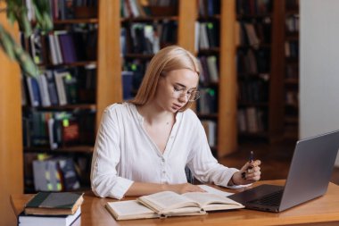 Ciddi derecede sıkı çalışan sarışın kız öğrenci webinar izliyor, çevrimiçi öğrenim kurslarını dinliyor, dizüstü bilgisayara bakıyor, üniversite kütüphanesindeki masada notlar yazıyor.