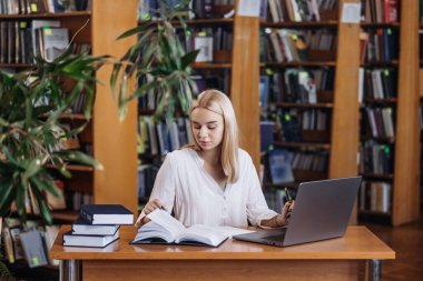 Ciddi derecede sıkı çalışan sarışın kız öğrenci webinar izliyor, çevrimiçi öğrenim kurslarını dinliyor, dizüstü bilgisayara bakıyor, üniversite kütüphanesindeki masada notlar yazıyor.