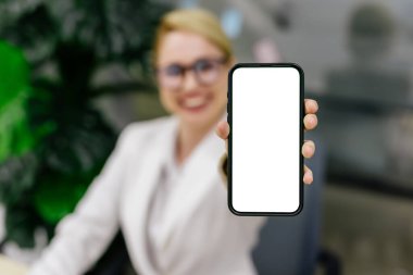 Modern bir ofiste, genç bir kadın elinde beyaz perdeyle bir telefon tutar. Dijital reklamları, etkileşimli içeriği veya tanıtım mesajlarını görüntülemek için ideal.