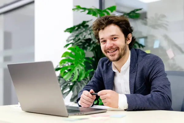 Karizmatik genç programcı, modern bir ofiste güven içinde oturuyor ve bilgisayarının yanındaki kameraya bakıyor. Teknoloji uzmanlarının temaları, modern çalışma alanları ve kariyer özgüveni için mükemmel..