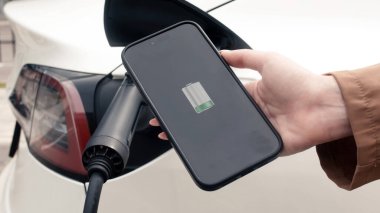 Elektrikli araba şarj edilirken telefonu kontrol eden kadının yakın görüntüsü. Modern elektrikli araçlar, teknoloji ve enerji kavramı.              
