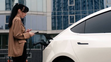 Genç bir kadın elektrikli arabayı şarj ediyor ve istasyonda telefon kullanıyor.