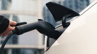 Elektrikli araba şarj olurken adam telefonu kontrol ediyor. Modern elektrikli araçlar, teknoloji ve enerji kavramı.              