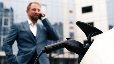 Elektrikli araba şarj olurken adam telefonu kontrol ediyor. Modern elektrikli araçlar, teknoloji ve enerji kavramı.              