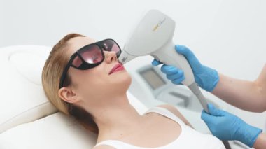 Modern bir klinikte kozmetik uzmanı lazerle saç aldırıyor. Genç bir kadın tüysüz ve pürüzsüz cilt için profesyonel tedavi görüyor.