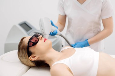 Modern bir klinikte kozmetik uzmanı lazerle saç aldırıyor. Genç bir kadın tüysüz ve pürüzsüz cilt için profesyonel tedavi görüyor.