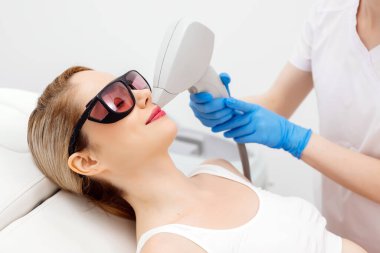 Modern bir klinikte kozmetik uzmanı lazerle saç aldırıyor. Genç bir kadın tüysüz ve pürüzsüz cilt için profesyonel tedavi görüyor.