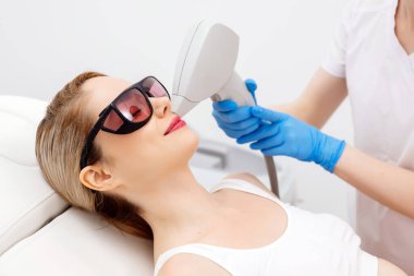 Modern bir klinikte kozmetik uzmanı lazerle saç aldırıyor. Genç bir kadın tüysüz ve pürüzsüz cilt için profesyonel tedavi görüyor.