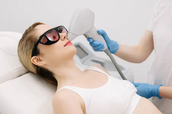 Modern bir klinikte kozmetik uzmanı lazerle saç aldırıyor. Genç bir kadın tüysüz ve pürüzsüz cilt için profesyonel tedavi görüyor.