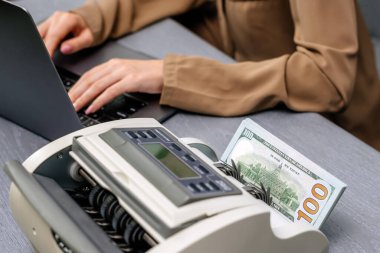 Elektronik para sayma makinesinde dizüstü bilgisayarı ve dolar banknotları olan kadın işçilere yakın çekim, finansal hizmetleri, nakit yönetimini veya döviz kurunu resmetmek için ideal.                           