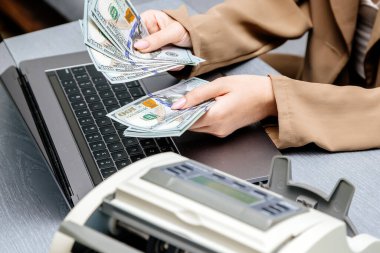 Elektronik para sayma makinesinde dizüstü bilgisayarı ve dolar banknotları olan kadın işçilere yakın çekim, finansal hizmetleri, nakit yönetimini veya döviz kurunu resmetmek için ideal.                           