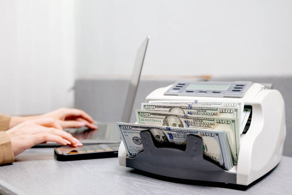 Крупный план женщины-работницы с ноутбуком и долларовыми купюрами в электронном автомате подсчета денег, идеально подходит для иллюстрации финансовых услуг, управления наличными или обмена валюты.                           