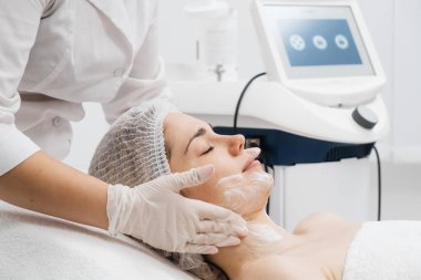 Kozmetik uzmanı modern bir güzellik kliniğinde gelişmiş cilt bakımı tekniklerini vurgulayan bir yüz kremi uyguluyor. Reklamınızda birinci sınıf spa tedavilerini sergilemek için ideal..     