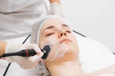 Kozmetik uzmanı modern bir klinikteki genç bir kadına radyasyon kaldırma uyguluyor. Bu gelişmiş işlem cilt bakımını sıkılaştırır ve gençleştirmeyi teşvik eder. Güzellik tedavileri için ideal..