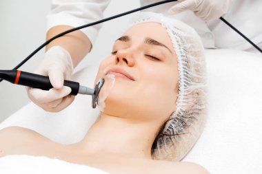 Kozmetik uzmanı modern bir klinikteki genç bir kadına radyasyon kaldırma uyguluyor. Bu gelişmiş işlem cilt bakımını sıkılaştırır ve gençleştirmeyi teşvik eder. Güzellik tedavileri için ideal..