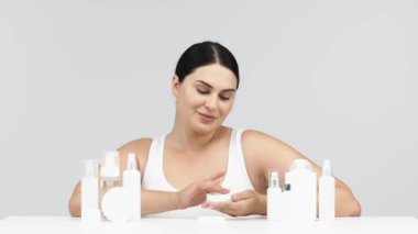 Gri arka planda kozmetik krem ve losyon şişeleriyle poz veren genç bir kadın.