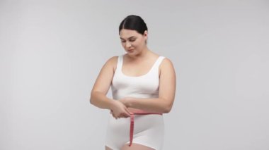 Kilolu bir kadın beyaz arka planda mezurayla poz veriyor.