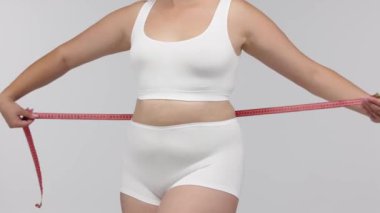 Vücut çalışmasından sonra vücudunu arka planda hafif bir şekilde ölçen bir kadın. Kilo verdirme, vites gösterme ve fitness hedeflerine doğru ilerlemeyi teşvik etmek için mükemmel..        