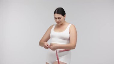 Kilolu bir kadın beyaz arka planda mezurayla poz veriyor.