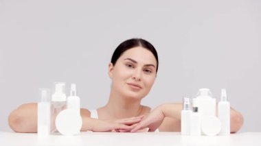 Gri arka planda kozmetik krem ve losyon şişeleriyle poz veren genç bir kadın.