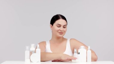 Gri arka planda kozmetik krem ve losyon şişeleriyle poz veren genç bir kadın.