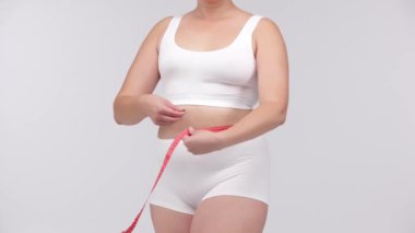 Vücut çalışmasından sonra vücudunu arka planda hafif bir şekilde ölçen bir kadın. Kilo verdirme, vites gösterme ve fitness hedeflerine doğru ilerlemeyi teşvik etmek için mükemmel..        