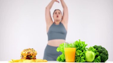 Fast food masada sağlıklı yiyeceğe karşı, arka planda egzersiz yapan genç kadın.