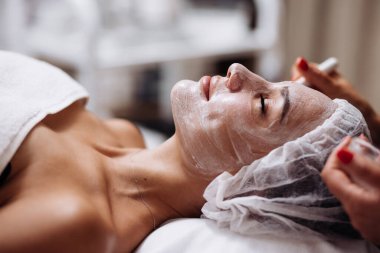 Bir kozmetolog modern bir güzellik kliniğinde cilt bakımı yöntemlerini vurgulayarak yüz kremi uyguluyor. Spa servisinin reklamını yapmak için mükemmel. Parlak tenli bir kadın. Makyaj yok