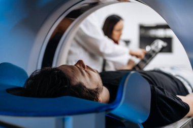 Modern hastanedeki MR makinesindeki genç adam ve arka plandaki teşhisi tartışan erkek ve kadın doktorlar.