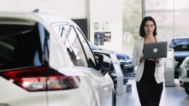 Kadın danışman, modern araba galerisindeki şık araçlarla çevrili dizüstü bilgisayardaki verileri inceliyor.       