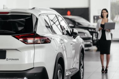 Kadın danışman, modern araba galerisindeki şık araçlarla çevrili dizüstü bilgisayardaki verileri inceliyor.       
