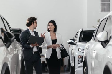 Kendine güvenen araba satıcısı modern araba galerisindeki alıcıyla yeni bir arabanın indirimlerini ve faydalarını tartışıyor.