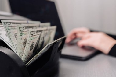 Bilgisayarın yanında para sayma makinesi olan tanınmayan bir kadın para bozdurmak ya da para yatırmak için yüz dolarlık banknotları sıralıyor. Finansal konsept, kredi veya volt değişimi.