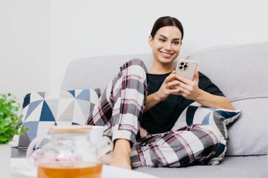 Genç ve güzel bir kadın, günlük giysiler içinde akıllı telefon kullanıyor ve evde kanepede dinlenirken gülümsüyor.
