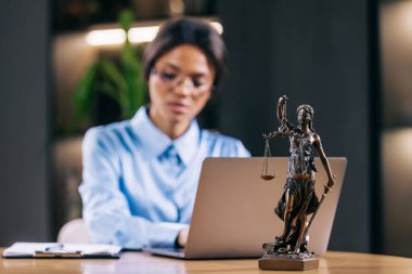 Dizüstü bilgisayarla çalışan konsantre genç avukat kadın ve masadaki tanrıça heykeli, seçici bir şekilde heykele odaklanmış.