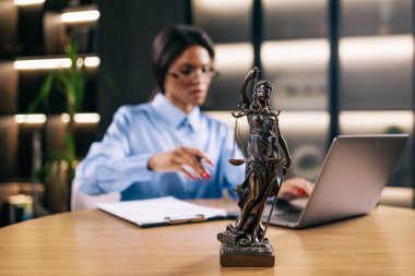 Dizüstü bilgisayarla çalışan konsantre genç avukat kadın ve masadaki tanrıça heykeli, seçici bir şekilde heykele odaklanmış.