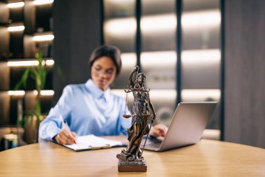 Dizüstü bilgisayarla çalışan konsantre genç avukat kadın ve masadaki tanrıça heykeli, seçici bir şekilde heykele odaklanmış.