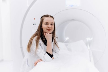 Hastanede MRI makinesinin yanında poz veren gülümseyen genç bayan doktor.