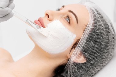 Profesyonel bir güzellik salonunda cilt bakımı sırasında genç bir kadının yakın çekimi. Kozmetik uzmanı kilden bir maske takıyor. Kozmetik, cilt bakımı ve cilt tazeleme kavramı.