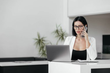 Genç iş kadını ofiste çalışırken telefonla konuşuyor ve laptop kullanıyor.