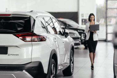 Resmi giyinen genç bayan yönetici yeni modern otomobille galeride dizüstü bilgisayarlı duruyor.