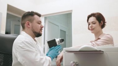 Bir dermatolog modern bir klinikte genç bir hastanın cildini gelişmiş ekipmanlarla inceler ve analiz eder. Deri araştırmaları, onkolojik deri hastalıklarının teşhisi.