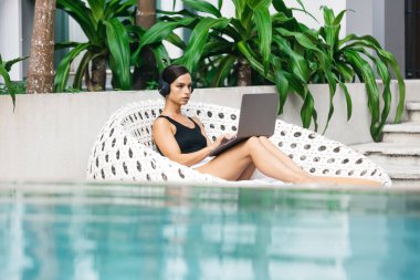 Kulaklıklı bir kadın tropikal ortamda çalışıyor ya da ders çalışıyor. Herhangi bir ortamda uzaktan çalışma, esneklik ve iş kavramı.