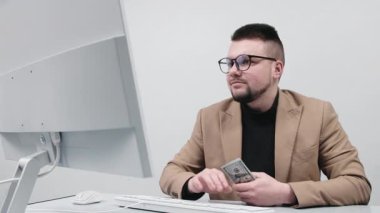 Gözlüklü genç bir işadamı modern bir ofiste elinde büyük miktarda nakit para tutuyor. Konsept, finansal bağımsızlığı ve iş hayatında başarıyı elde etmenin hissini aktarmak..