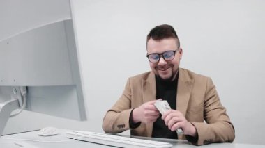 Gözlüklü genç bir işadamı modern bir ofiste elinde büyük miktarda nakit para tutuyor. Konsept, finansal bağımsızlığı ve iş hayatında başarıyı elde etmenin hissini aktarmak..