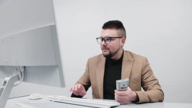 Gözlüklü genç bir işadamı modern bir ofiste elinde büyük miktarda nakit para tutuyor. Konsept, finansal bağımsızlığı ve iş hayatında başarıyı elde etmenin hissini aktarmak..