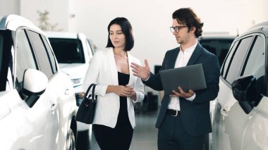 Araba satıcısı potansiyel alıcılarla görüşür, çeşitli arabalarla dolu parlak bir galeride araç özelliklerini tartışır, müşteri hizmetleri deneyimini ön plana çıkarır