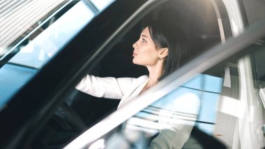 Beyaz takım elbiseli bir kadın lüks bir arabanın içini incelerken çağdaş tasarım ve entelektüelliği ön plana çıkarıyor.